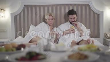 放松的高加索男人和女人躺在床上喝咖啡，喝果汁，说话。 入住酒店休息的快乐成年夫妇